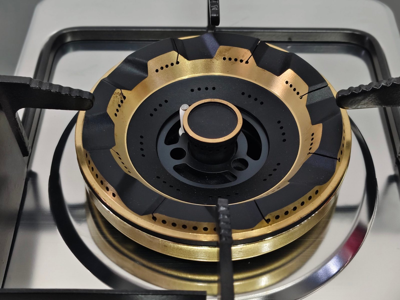 A close-up of the GCS-71306-B-04.jpg built-in gas hob shows a modern black and gold burner with multiple flame ports, metallic supports, and a reflective stainless steel surface.