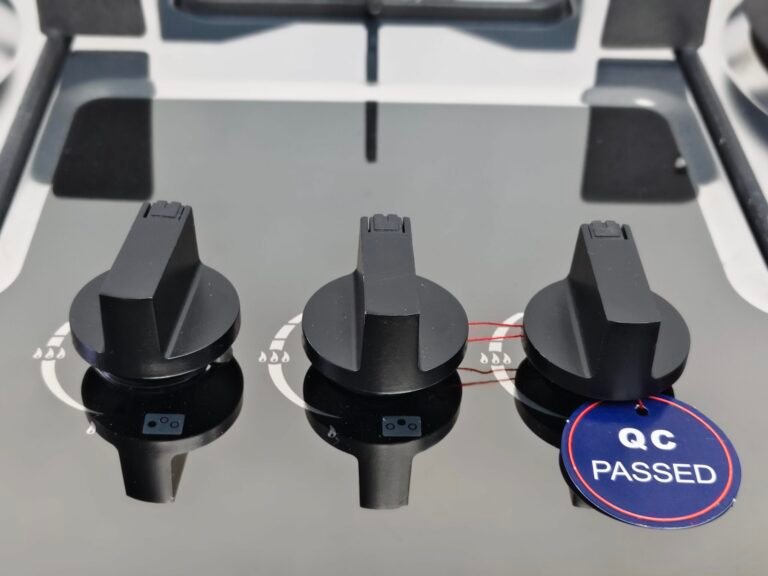 Three black stove control knobs are mounted on the shiny black surface of the GCS-73301-B-02 gas hob with three burners. The rightmost knob has a blue "QC PASSED" tag, and their reflections appear on the hob’s built-in surface.