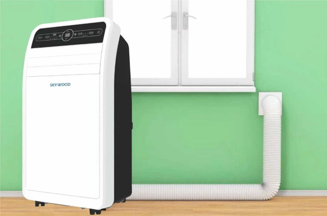 The SKP-1221-03.webp portable inverter air conditioner sits on a wooden floor against a green wall, with its hose connected to a window vent beneath a closed window.