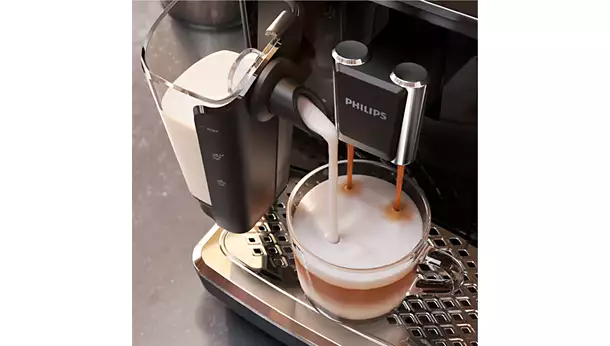 The vrs_5400-09-ac.webp fully automatic coffee machine pours steamed milk and espresso into a clear glass, creating a beautifully layered coffee drink on a metal drip tray.